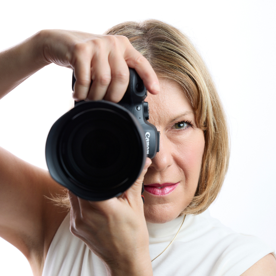 NYC Photographer, Anne Vandycke, holding a Canon camera lens close to her eye against a white background.