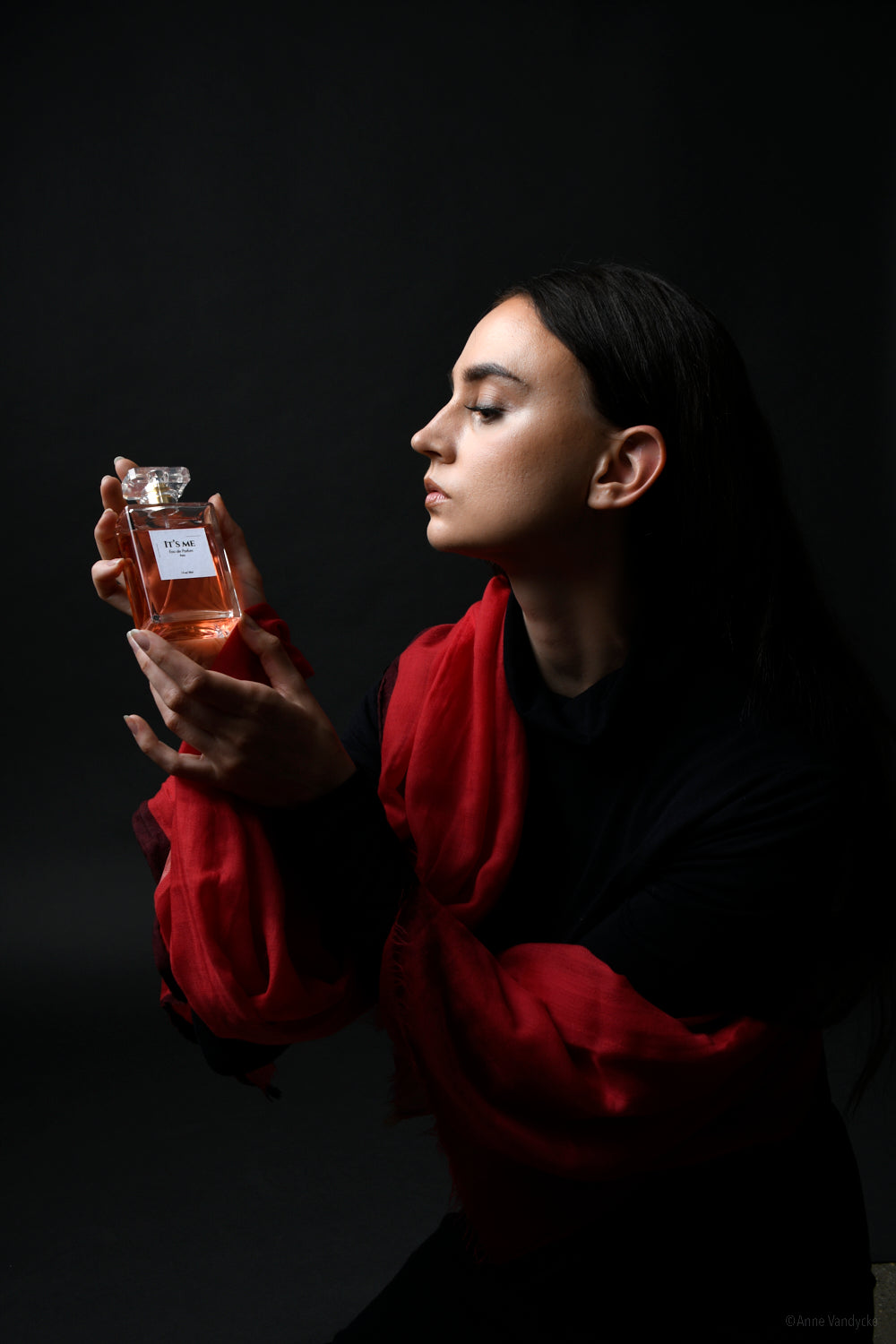 Woman holding a perfume bottle against a dark background wearing red scarf. Photo styled by New York Photographer, Anne Vandycke.