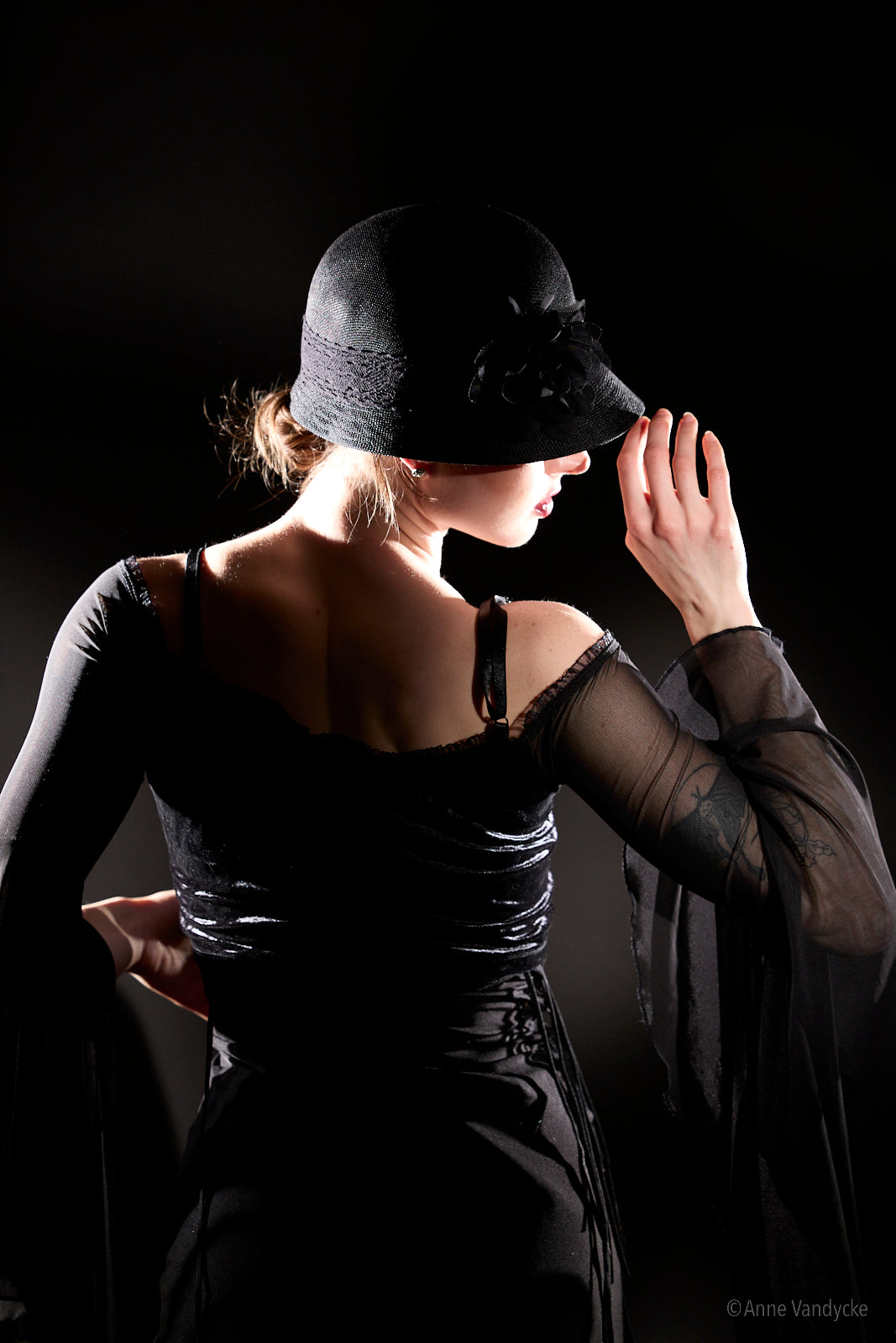 Woman in a black dress and hat against a dark background. Photo posed and taken by NY photographer, Anne Vandycke.