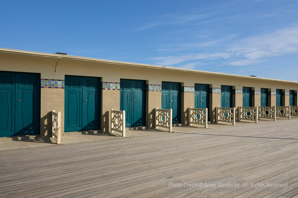 Row of beach houses with blue doors on a wooden deck under a clear blue sky. Architecture photography by Anne Vandycke.