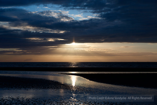 Sunset over a beach with dark clouds and reflective water. Sunset photo taken by New York Photographer, Anne Vandyke.