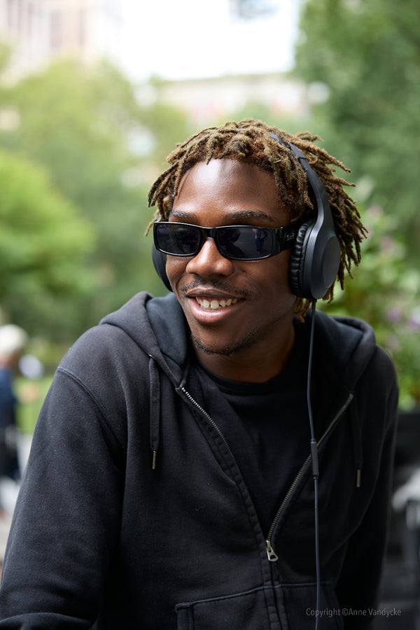 Man wearing sunglasses and headphones with a blurred outdoor background. Candid New York photo by Anne Vandycke.