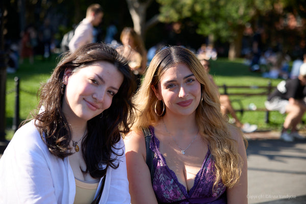 Two women sitting together in a park with people and trees in the background. New York City Photoshoot by Anne Vandycke.
