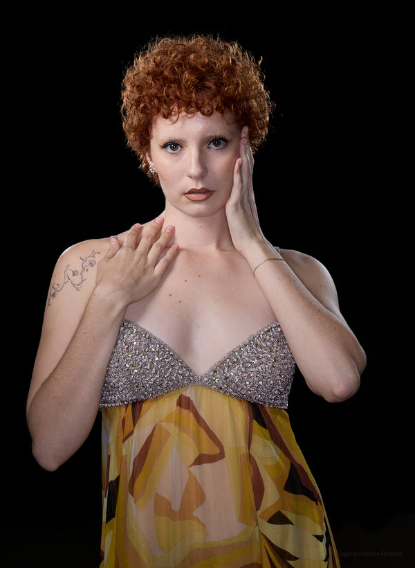 Woman with red hair wearing a silver top and yellow patterned dress against a black background. Posed studio photo by Anne Vandycke, NY photographer.