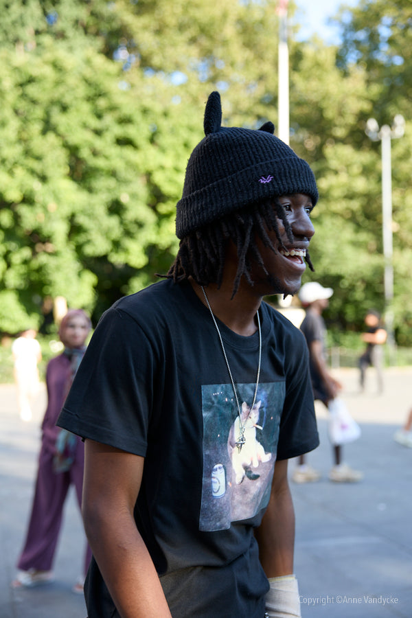 Person wearing a black beanie and t-shirt with a graphic design, standing outdoors. Candid Shot by New York photographer, Anne Vandycke.