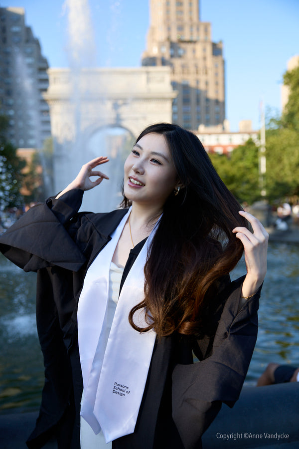 Woman in graduation gown with a white stole standing by a fountain with buildings in the background. Candid Photography by New York Photographer, Anne Vandycke.