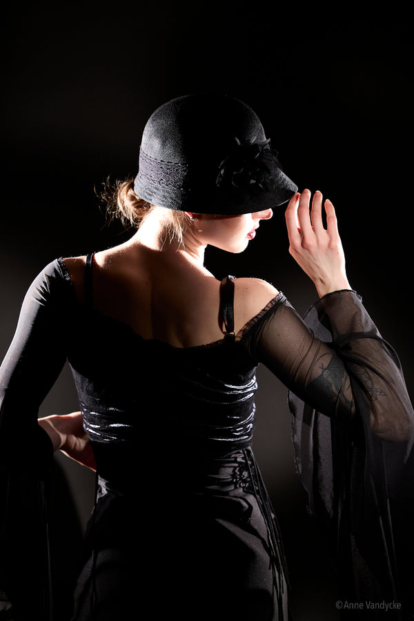 Woman in a black dress and hat against a dark background. Photo posed and taken by NY photographer, Anne Vandycke.