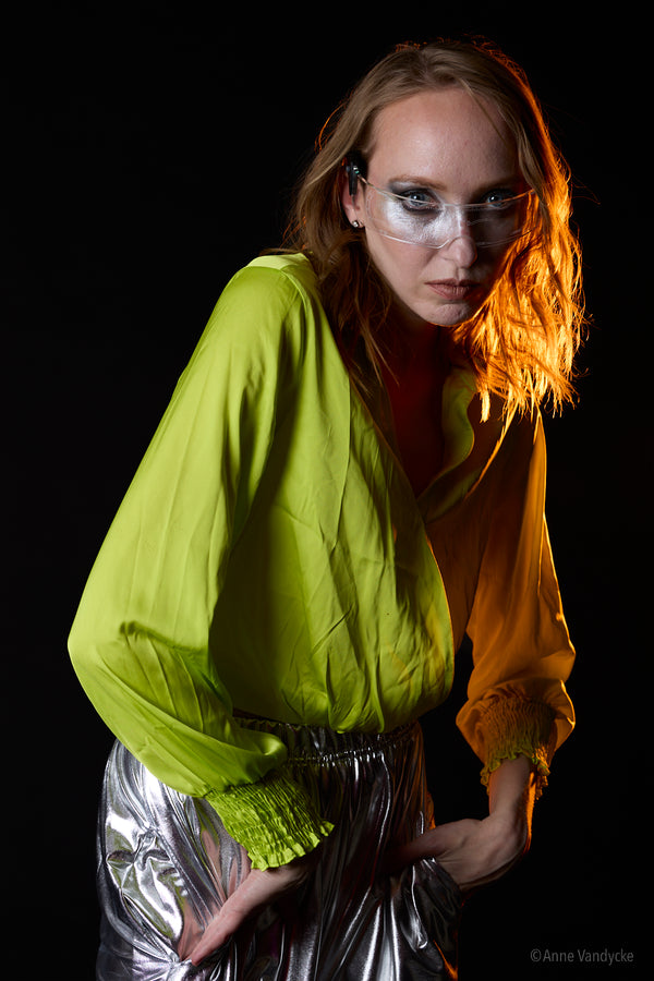 Woman wearing a bright green top and silver pants against a black background. Studio photography by Anne Vandycke.