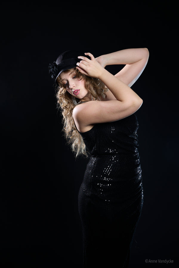 Woman in a black sequin dress and hat against a black background. Photo taken by New York photographer Anne Vandycke.