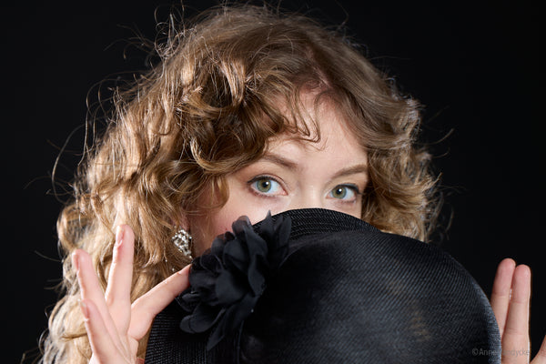 Woman with curly hair holding a black hat in front of her face against a dark background. Studio photo by Anne Vandycke. 