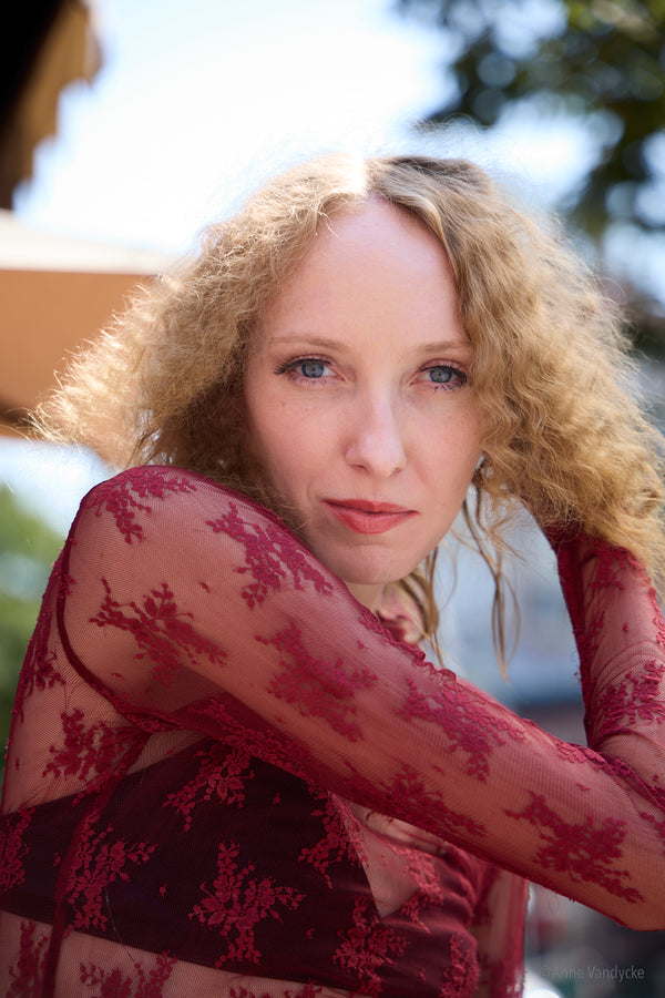 Woman with curly hair wearing a red floral top with a blurred outdoor background. Outdoor New York photos by Anne Vandycke.