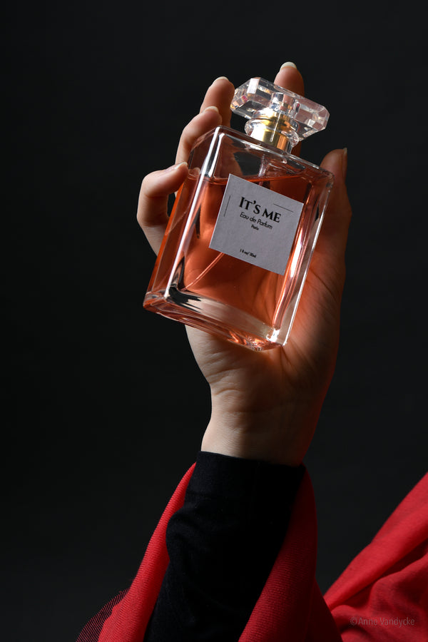 Hand holding a rectangle perfume bottle labeled 'It's Me' against a dark background. Photo styled and shot by New York photographer, Anne Vandycke.