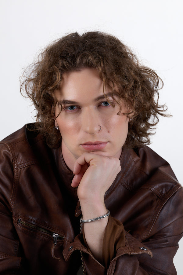 Man with curly hair wearing a brown leather jacket against a white background. Studio photos by New York photographer, Anne Vandycke/Copyright 2026 ©Anne Vandycke, All rights reserved. 