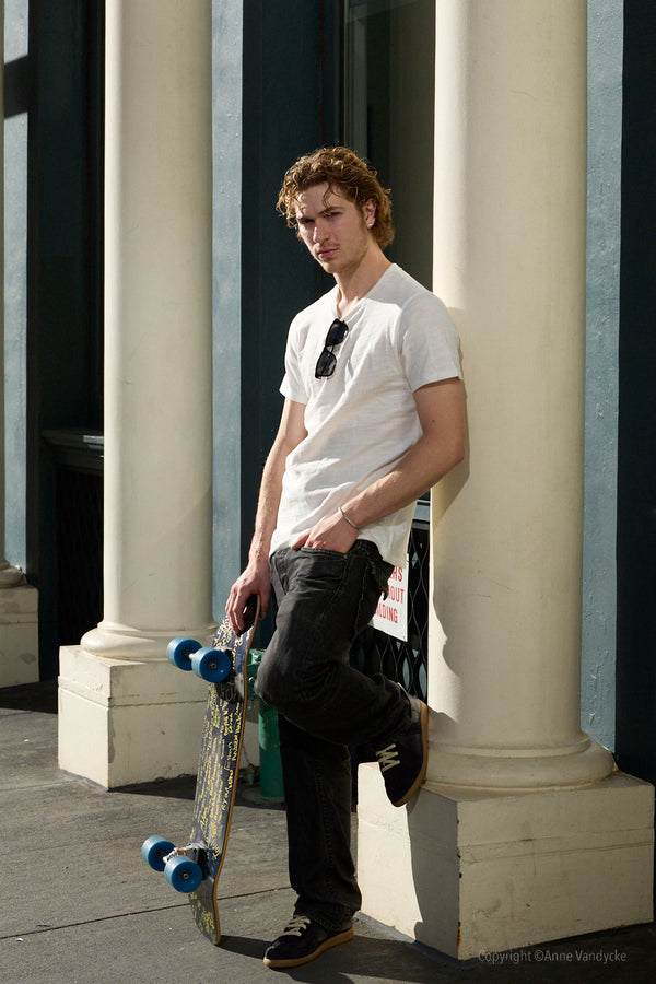 Man holding a skateboard leaning against a columned building. New York Photography by Anne Vandycke.