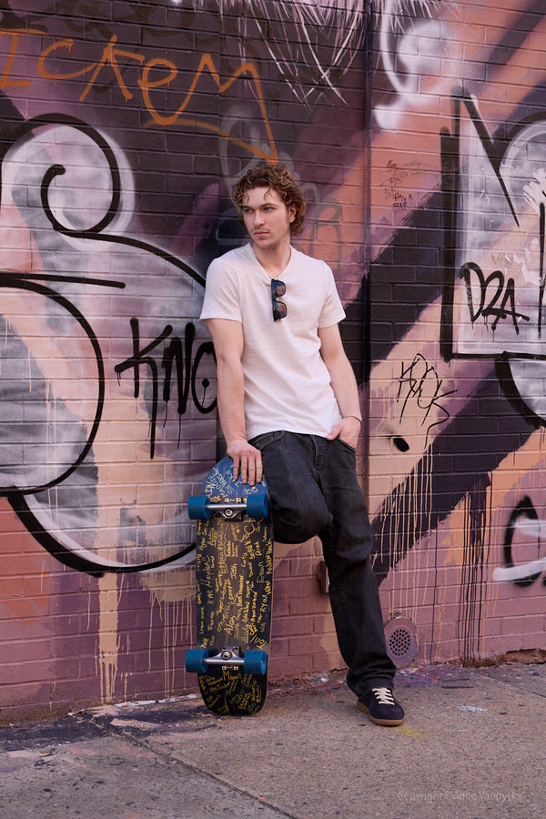Man holding a skateboard against a graffiti-covered wall. Photo by NY photographer, Anne Vandycke.