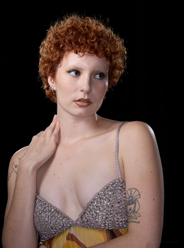 Woman with curly red hair wearing a sparkly top against a black background. New York City Photoshoot by Anne Vandycke.