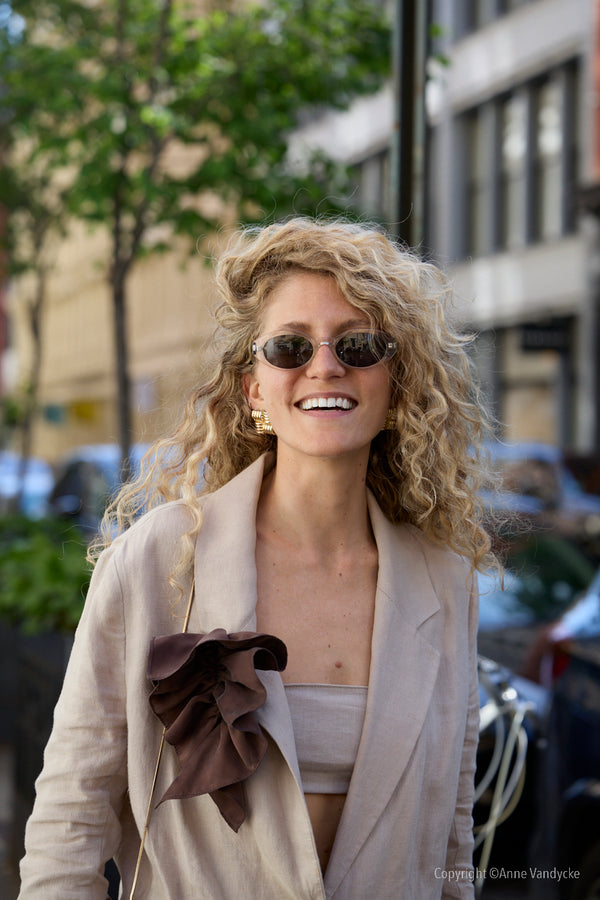 Woman with curly hair wearing sunglasses and a beige coat in an urban setting. Candid Shots Photography by Anne Vandycke.
